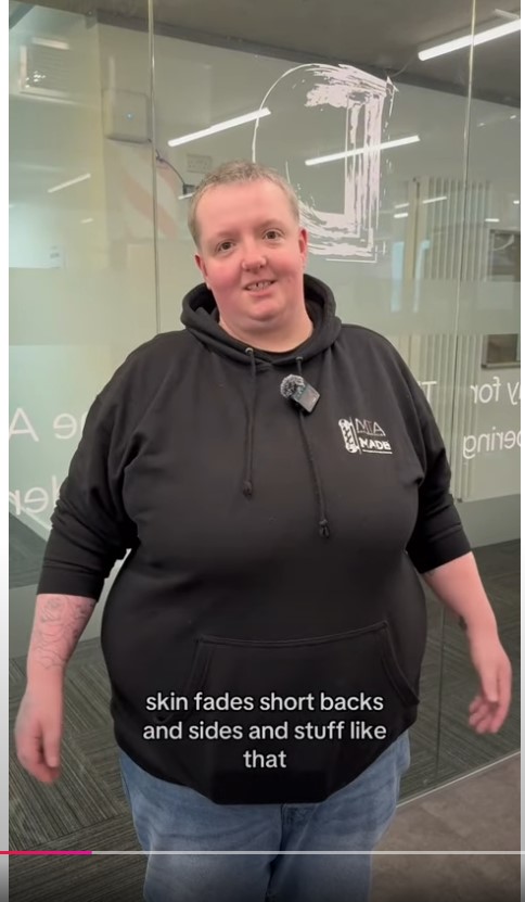 A barbering student in a black hoodie discussing skin fades and short back and sides in a training facility.