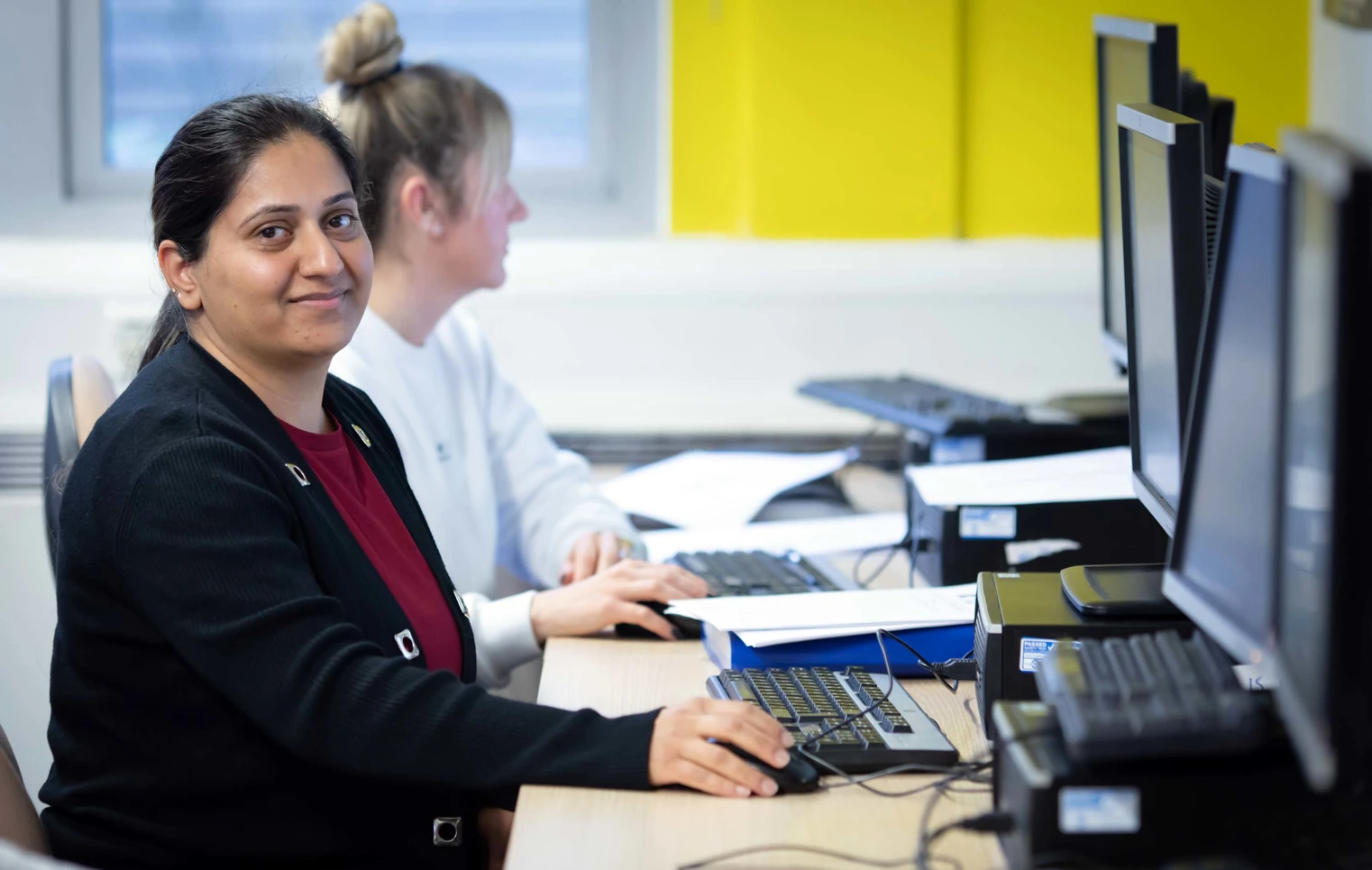 Student in a black cardigan sitting at a computer workstation, smiling while working on a task in a business and admin class. Student in a black cardigan sitting at a computer workstation, smiling while working on a task in a business and admin class.