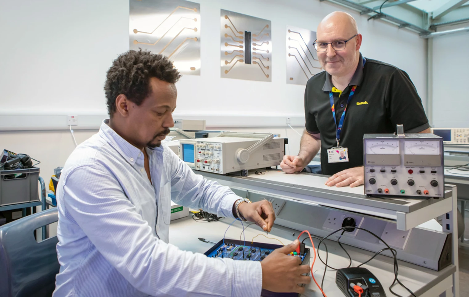 A lecturer and a student in an electronics lab working on a circuit board experiment. The lecturer is smiling while overseeing the student’s work. A lecturer and a student in an electronics lab working on a circuit board experiment. The lecturer is smiling while overseeing the student’s work.