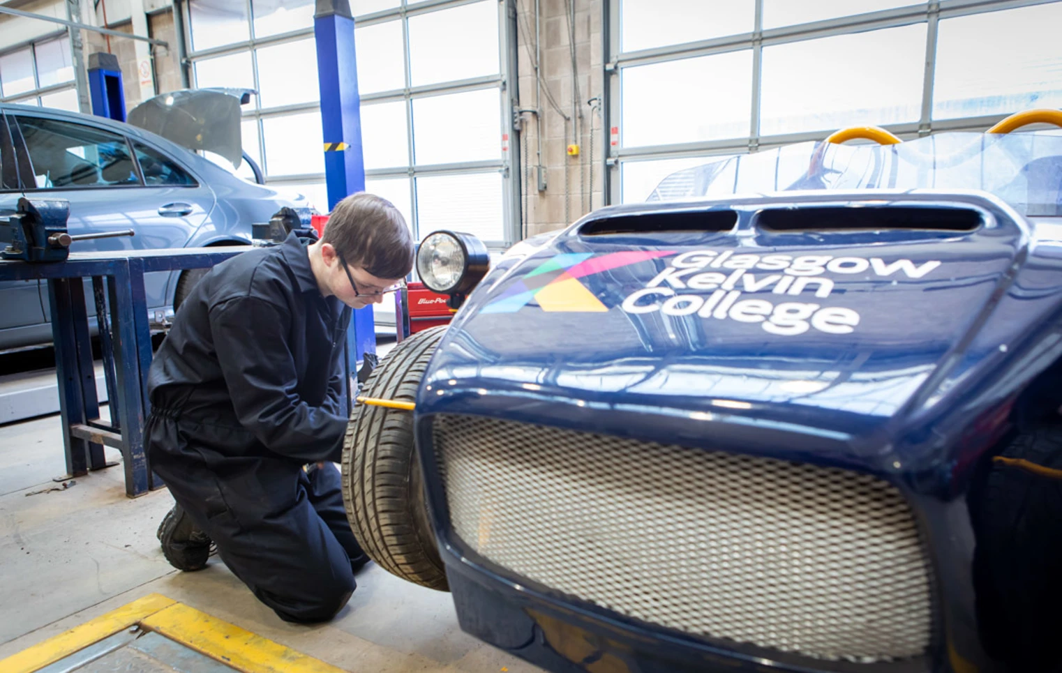Student working on the front wheel of a Glasgow Kelvin College-branded kit car, checking its alignment and assembly. Student working on the front wheel of a Glasgow Kelvin College-branded kit car, checking its alignment and assembly.