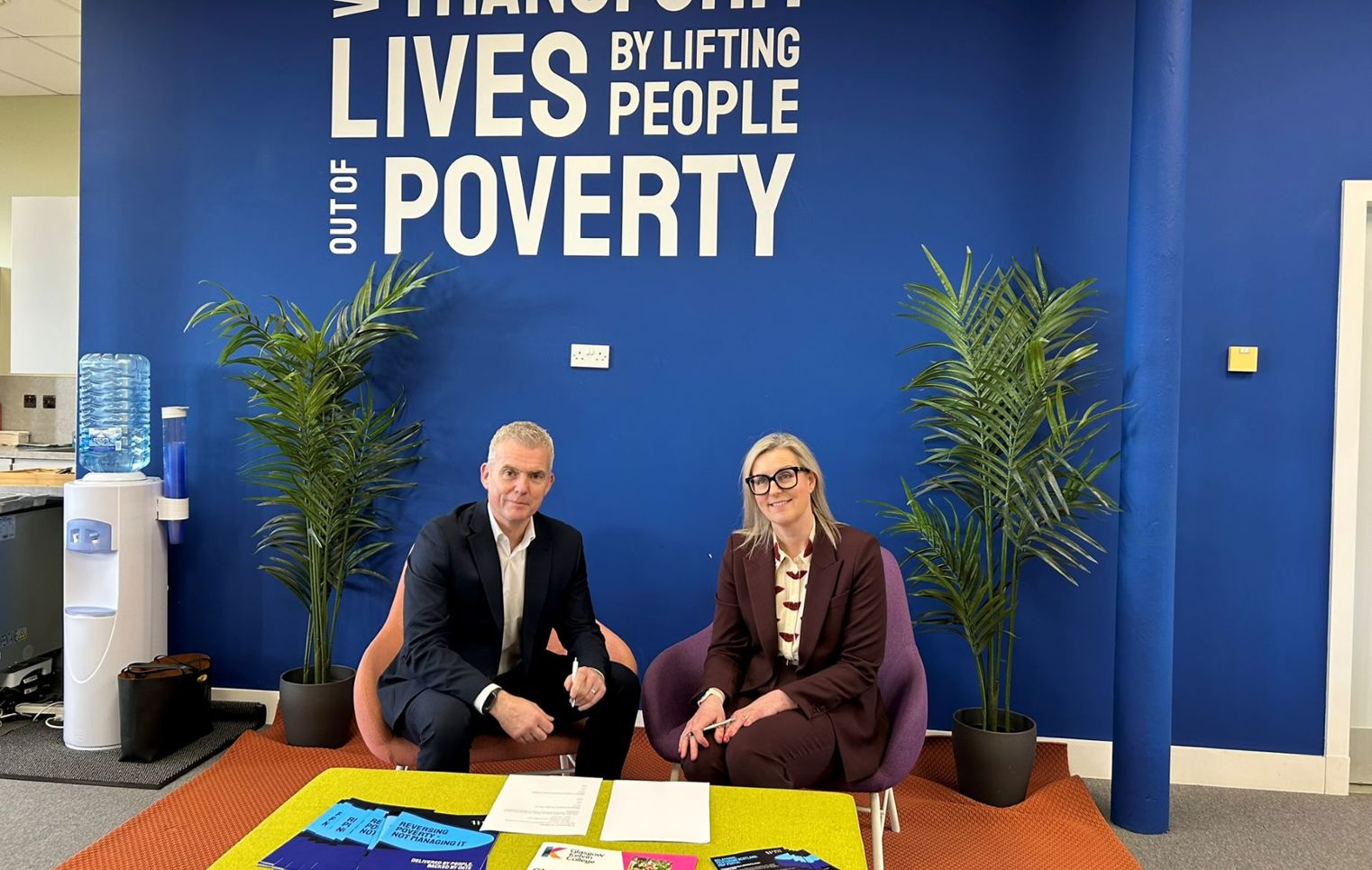 Two professionals seated beneath a “We Transform Lives by Lifting People Out of Poverty” wall, marking a partnership agreement.