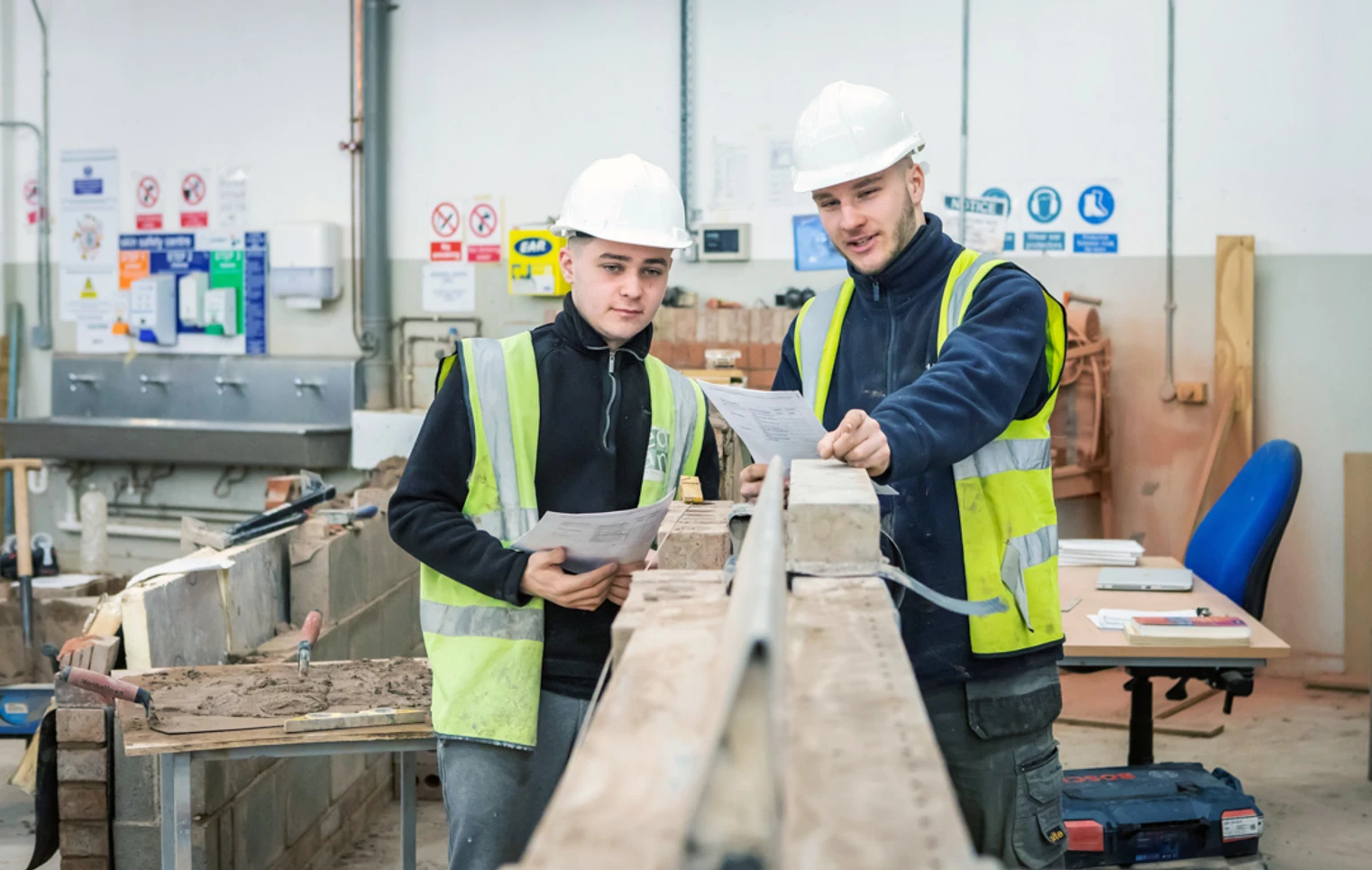 Two brickwork students in safety gear reviewing plans together, standing beside a brick wall and discussing their next steps in a practical training workshop. Two brickwork students in safety gear reviewing plans together, standing beside a brick wall and discussing their next steps in a practical training workshop.