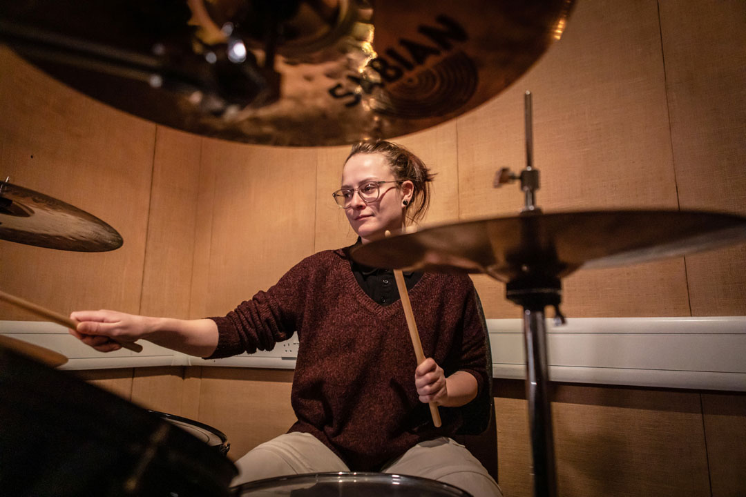A young drummer with glasses and a burgundy sweater, playing a drum kit surrounded by cymbals.