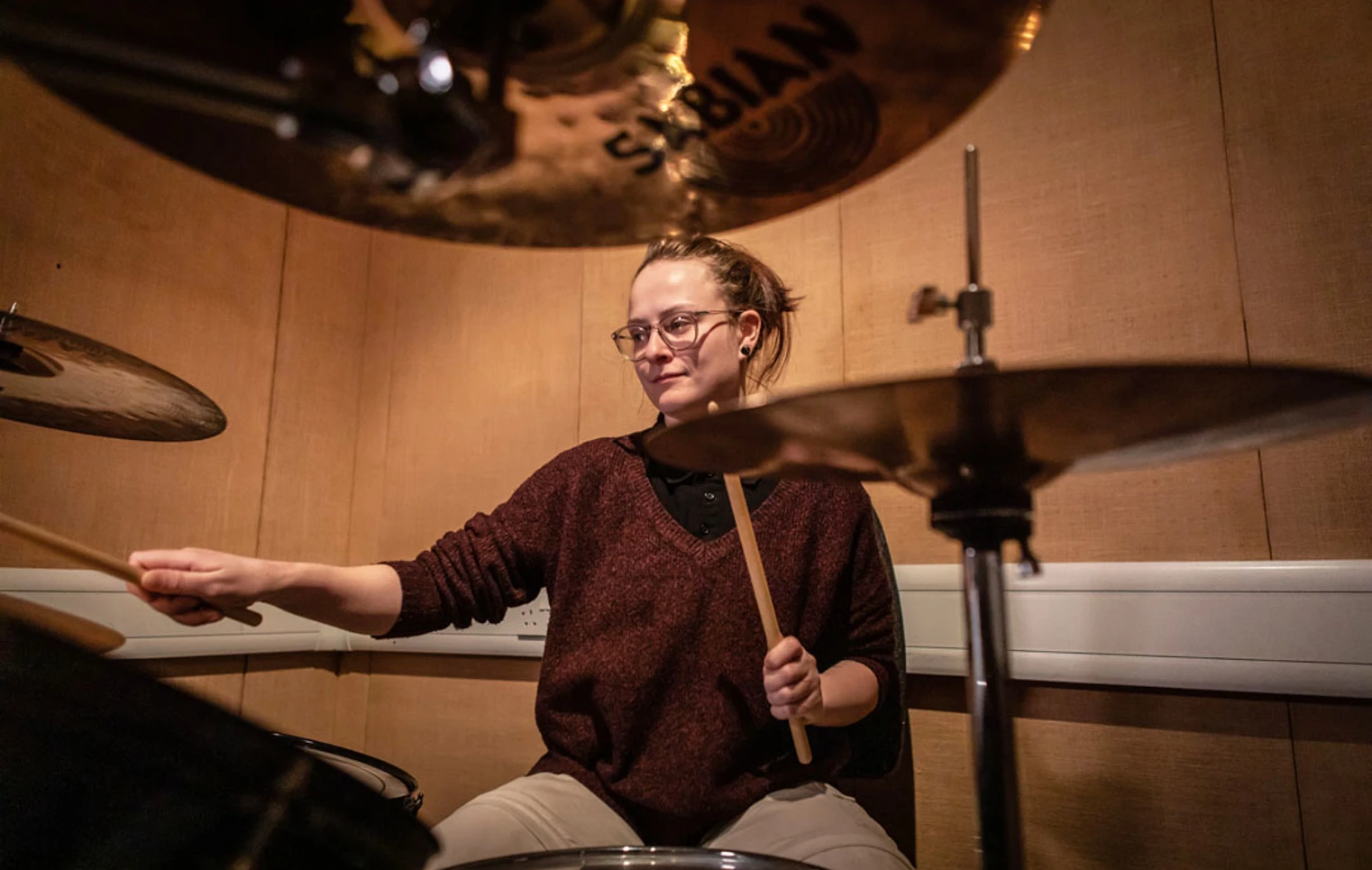 A young drummer with glasses and a burgundy sweater, playing a drum kit surrounded by cymbals. A young drummer with glasses and a burgundy sweater, playing a drum kit surrounded by cymbals.