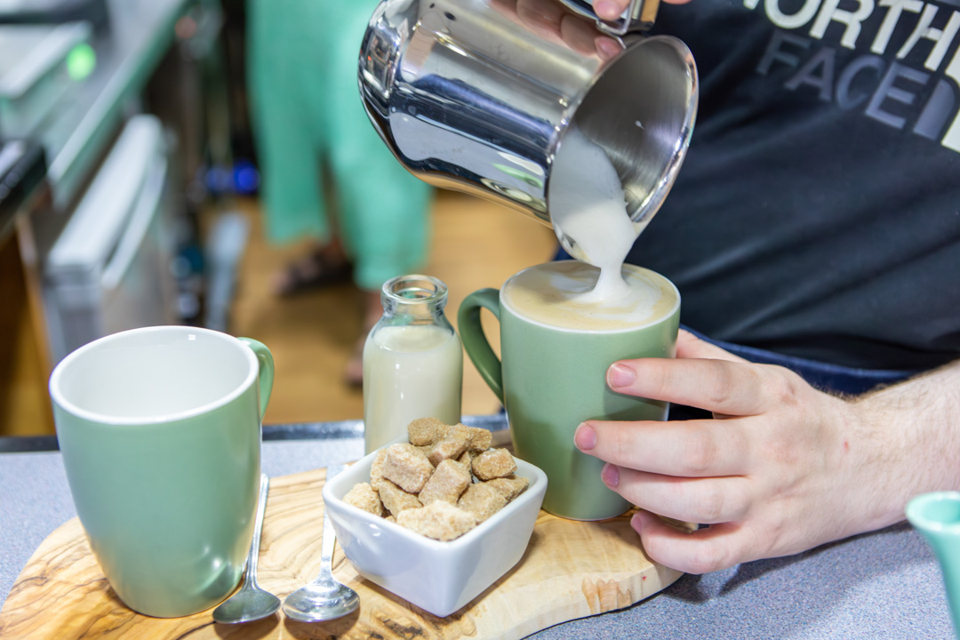 Close-up of a barista student pouring steamed milk into a cup of coffee on a wooden serving tray.
