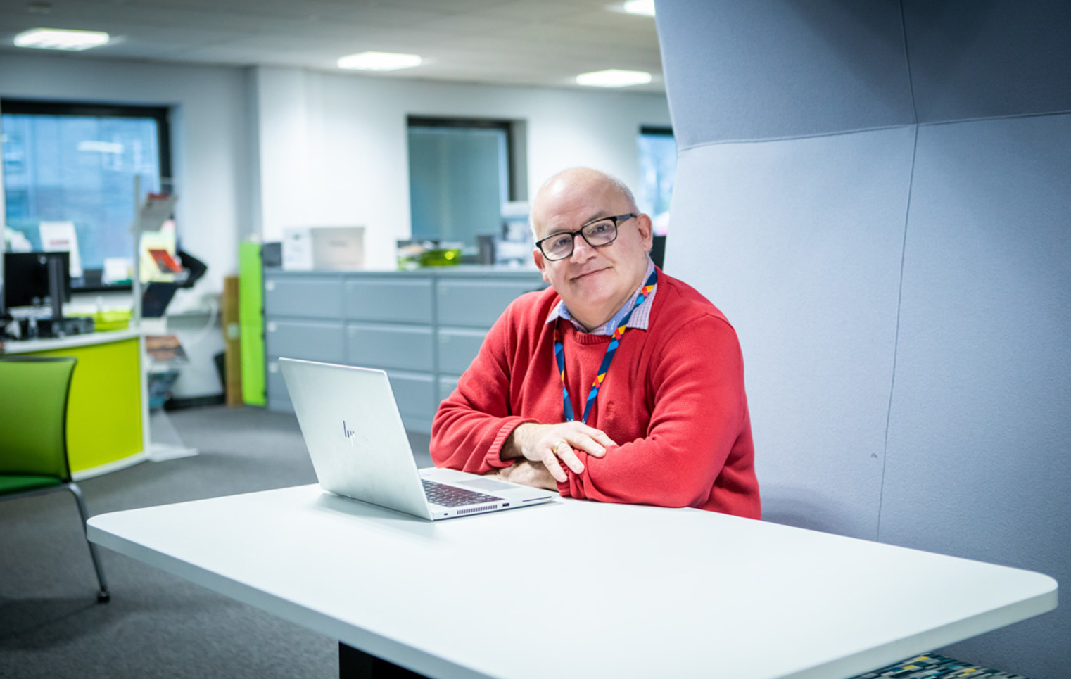 Smiling staff member in a red jumper sitting at a desk with a laptop in a modern office setting at Glasgow Kelvin College