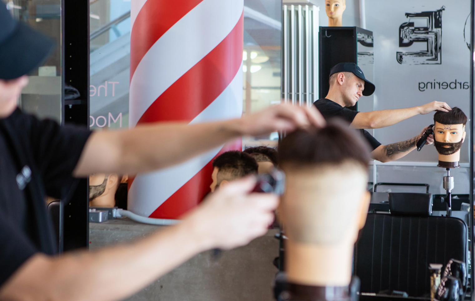 A barbering student using a spray bottle on a mannequin head while another student trims a different mannequin in the background.