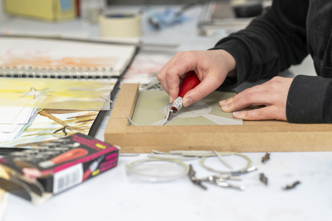 Close-up of hands carving a design into a printmaking block on a desk, surrounded by sketches and art supplies