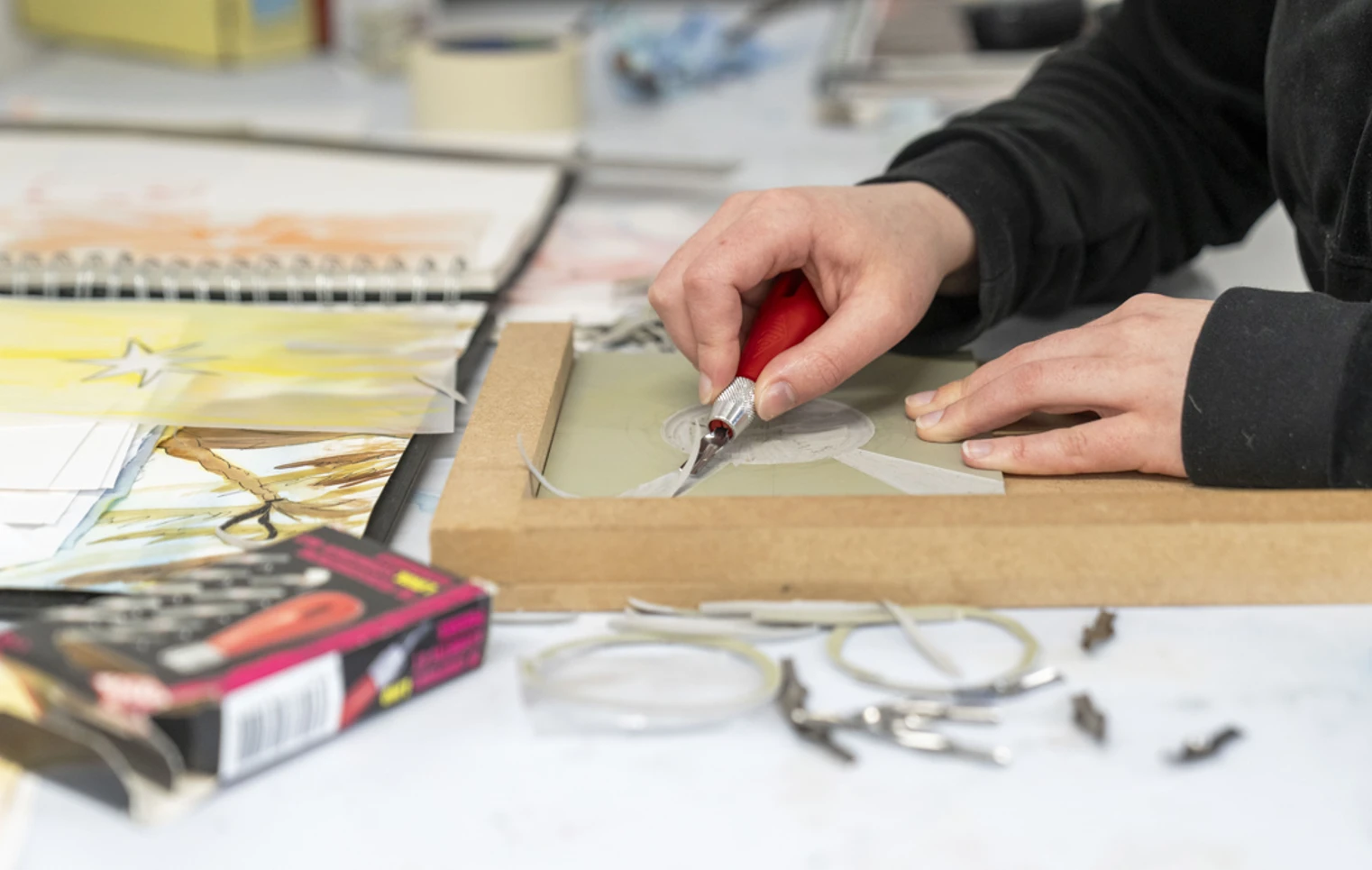 Close-up of hands carving a design into a printmaking block on a desk, surrounded by sketches and art supplies Close-up of hands carving a design into a printmaking block on a desk, surrounded by sketches and art supplies