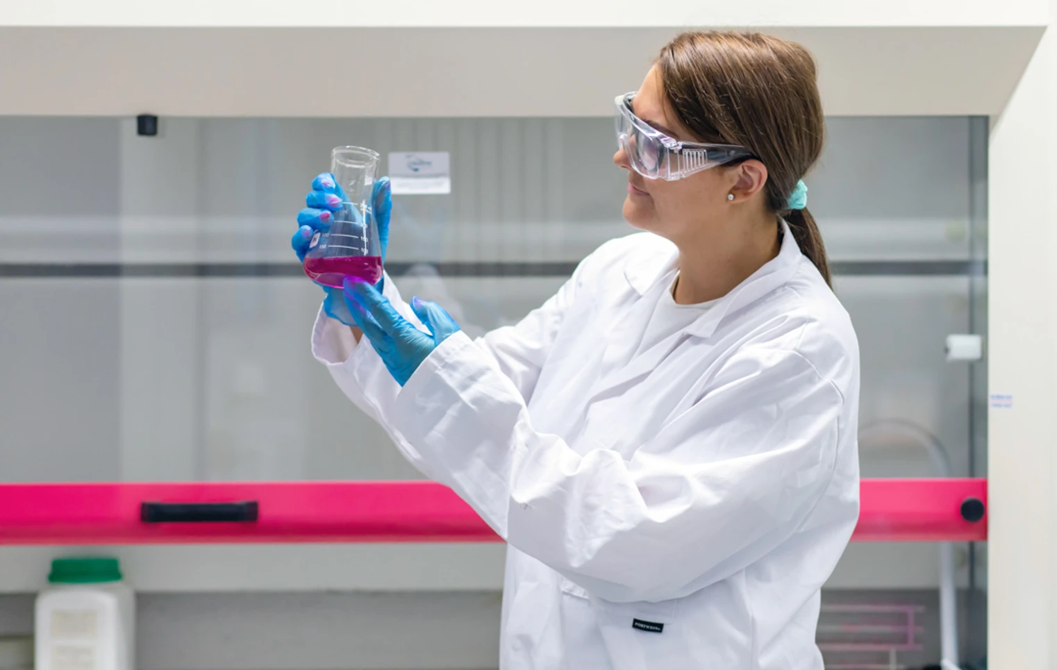 Student in a lab coat studying a beaker of pink liquid under bright lighting in a science lab. Student in a lab coat studying a beaker of pink liquid under bright lighting in a science lab.