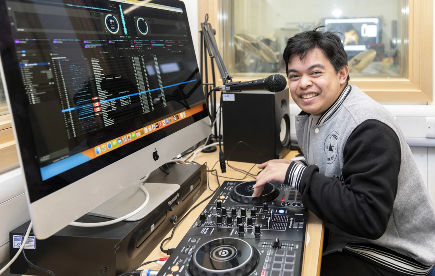 Young man smiling while using DJ equipment and a microphone, with a computer screen displaying music software. Young man smiling while using DJ equipment and a microphone, with a computer screen displaying music software.