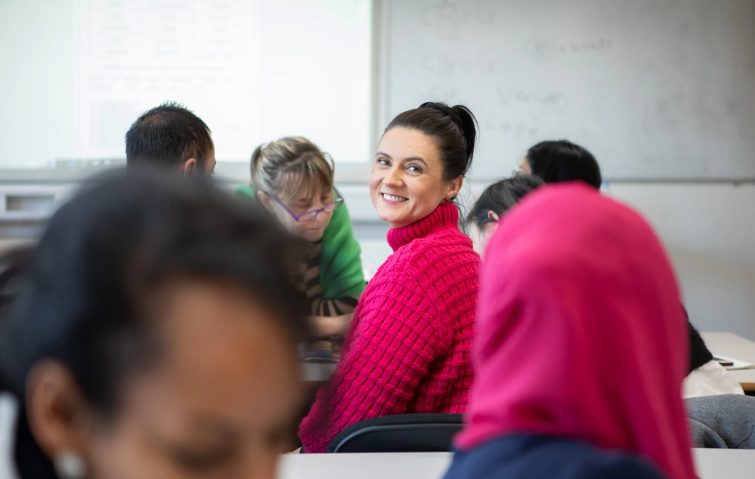 A woman in a bright pink sweater smiles at the camera while sitting in a classroom with other students working in the background.  A woman in a bright pink sweater smiles at the camera while sitting in a classroom with other students working in the background.