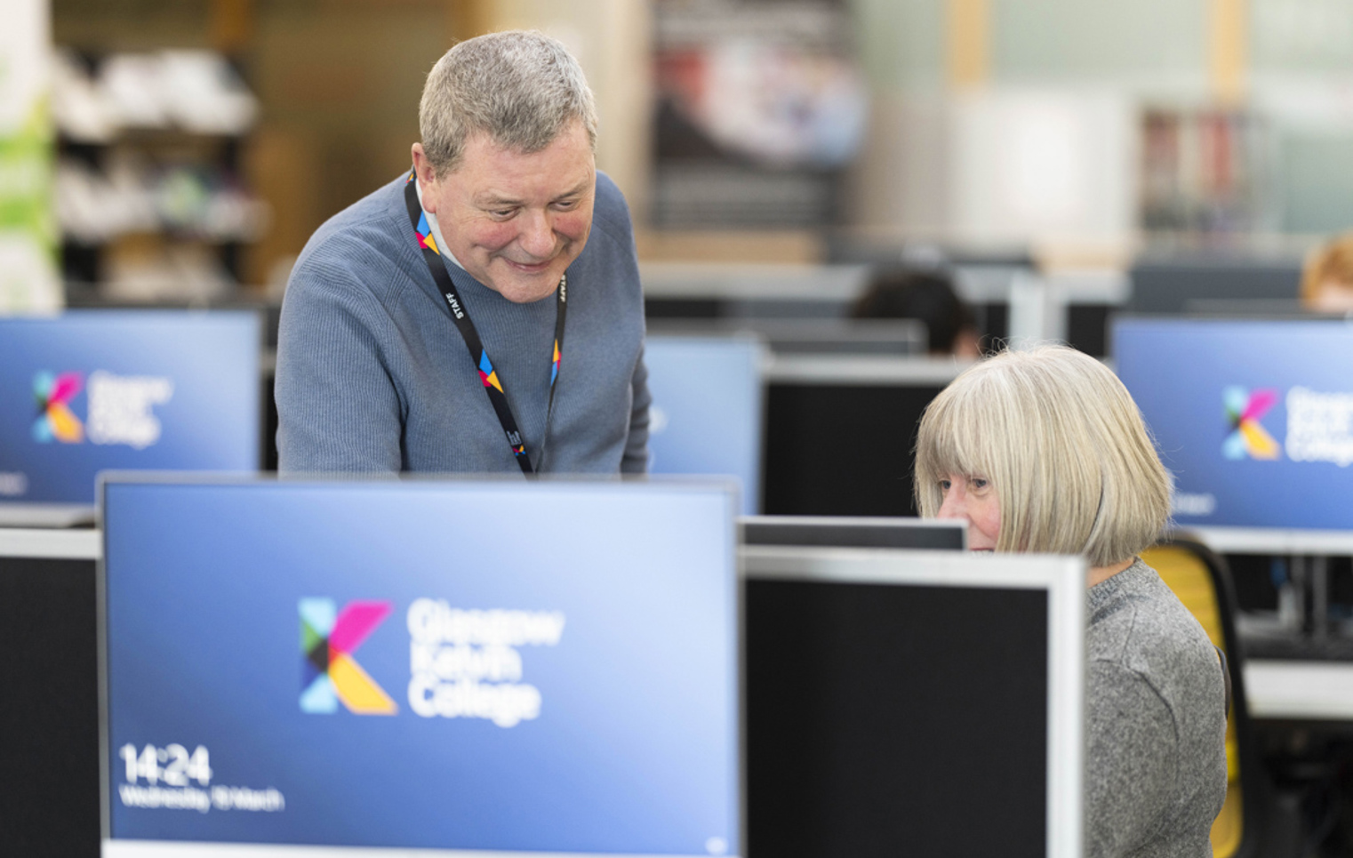 Staff member supporting a student in the computer suite at Glasgow Kelvin College, with college branding visible on screens.